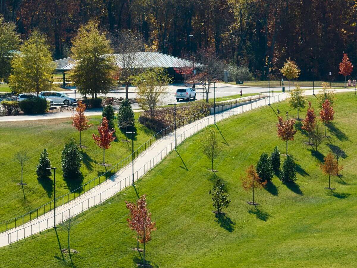 A fenced paved path moving leading down grassy a hill from a parking lot