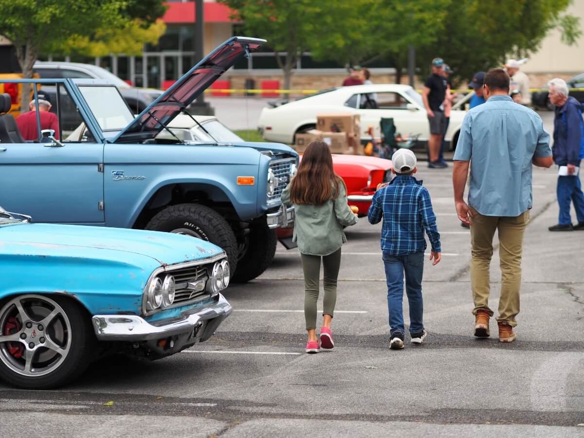A man and two children walking through a parking lot looking at vintage cars there for a car show