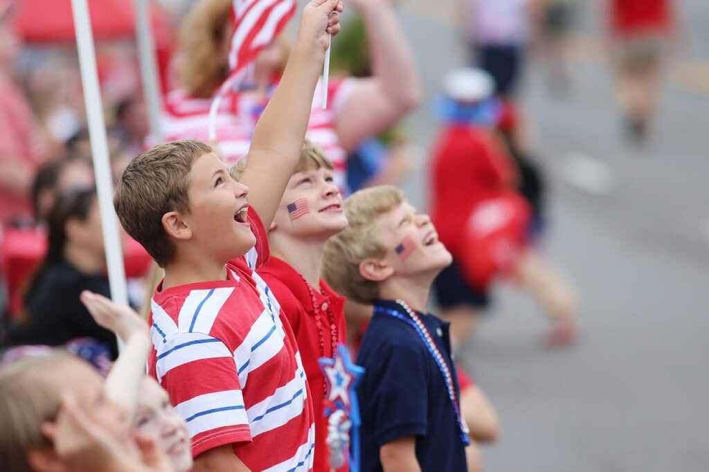 A few patriotic children wearing red, white, and blue staring up at something and waving an American flag